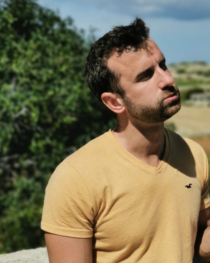 Marco Diversi outdoors in a mustard yellow t-shirt, looking up and to the side, with green trees and a blue sky behind him